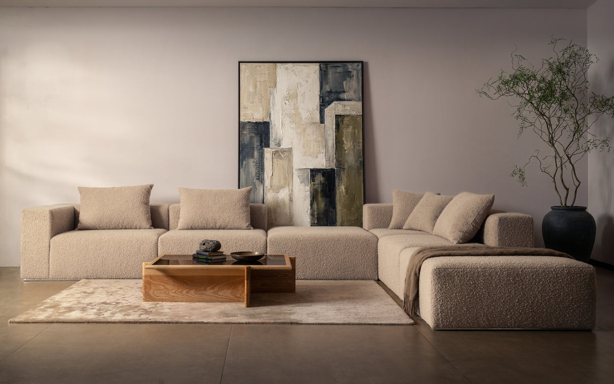 A modern beige sectional sofa with matching throw pillows in a minimalist living room setting, styled with a wooden coffee table, textured rug, and a large abstract wall art piece.