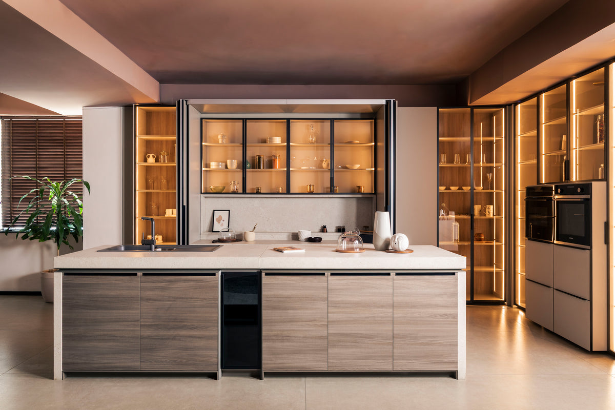 A modern kitchen island with light wood grain cabinets and a white countertop, set within a contemporary kitchen featuring warm ambient lighting, glass-front cabinets, and built-in appliances.