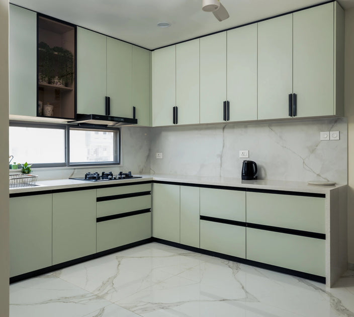 A modern kitchen interior with pale sage green cabinets, black handles, white marble-look countertops and backsplash, and a white marble floor. A built-in gas stove, sink, and window are visible, along with a small black appliance on the counter.10