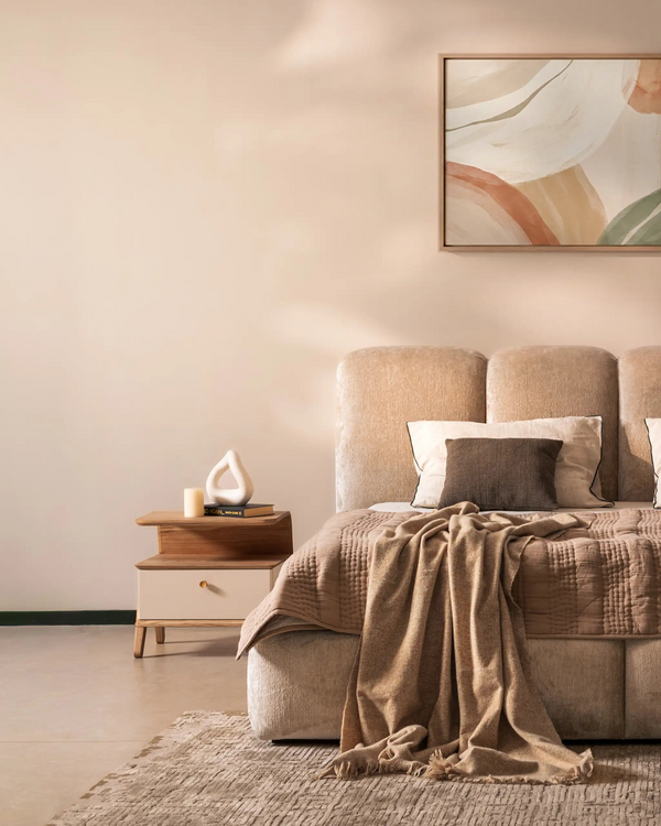 A styled bedroom scene featuring a beige upholstered bed with textured bedding and pillows, a matching beige throw blanket draped over the foot of the bed, a small wooden nightstand with a white vase, and a framed abstract painting on the wall.